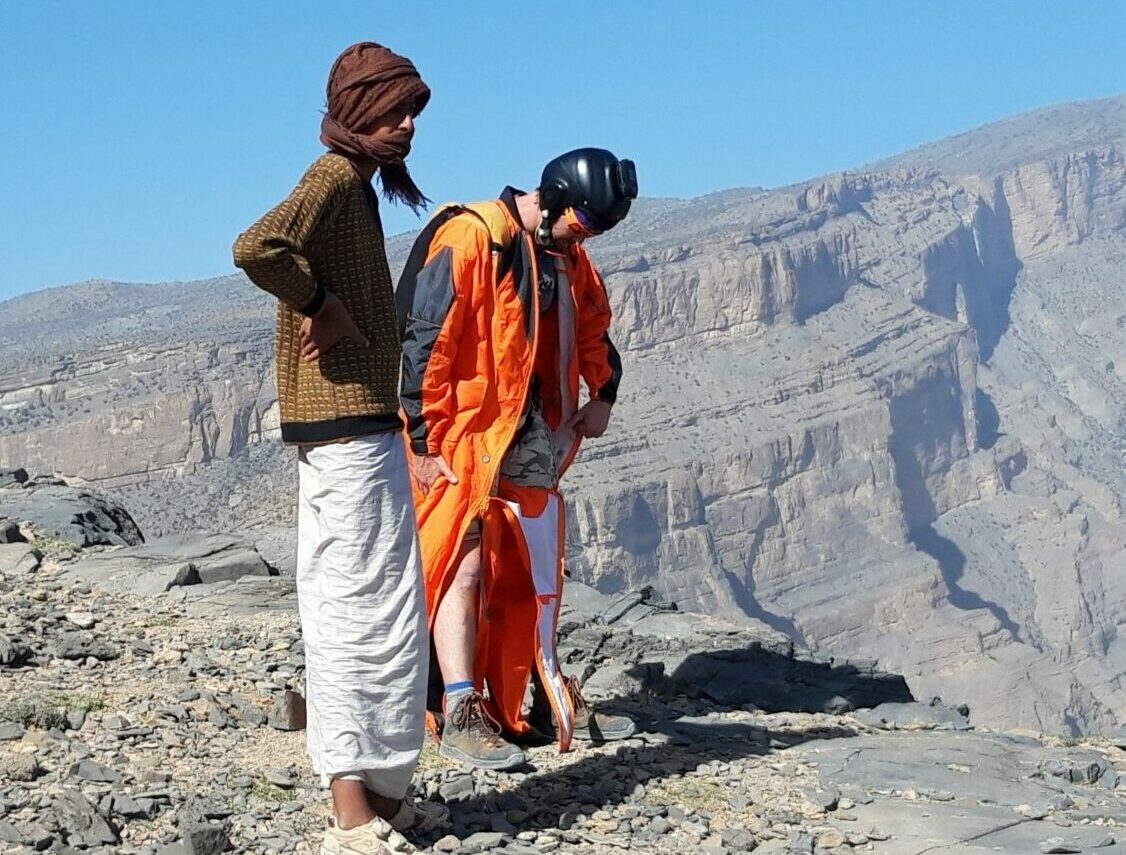 A base jumper wearing a wingsuit and an indigenous in traditionnal clothes stand together on the edge of a cliff in Oman.