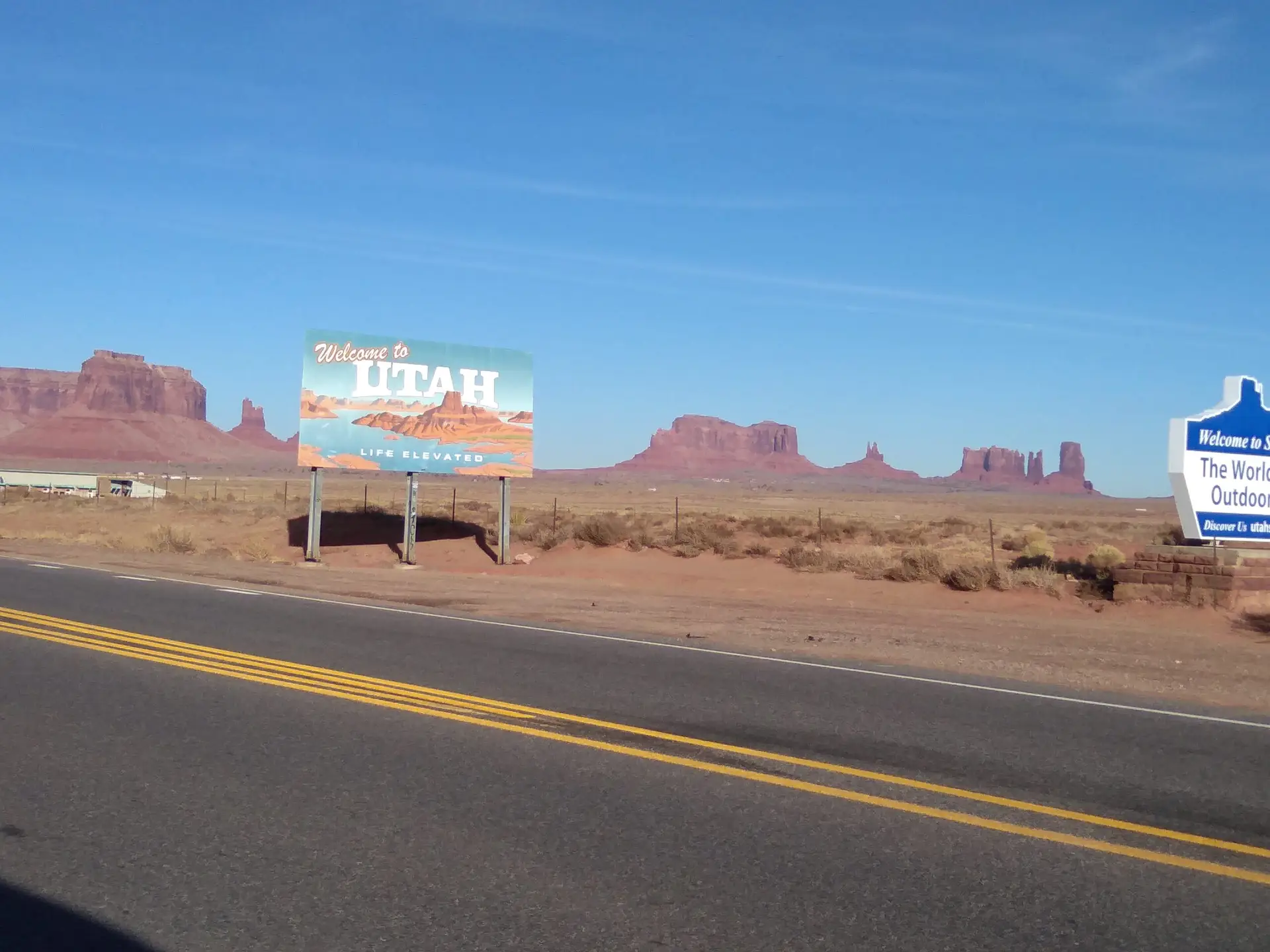 Au bord d'un route, panneau welcome to Utah avec en arrière-plan des mesas, formations géologiques de roche de sable.