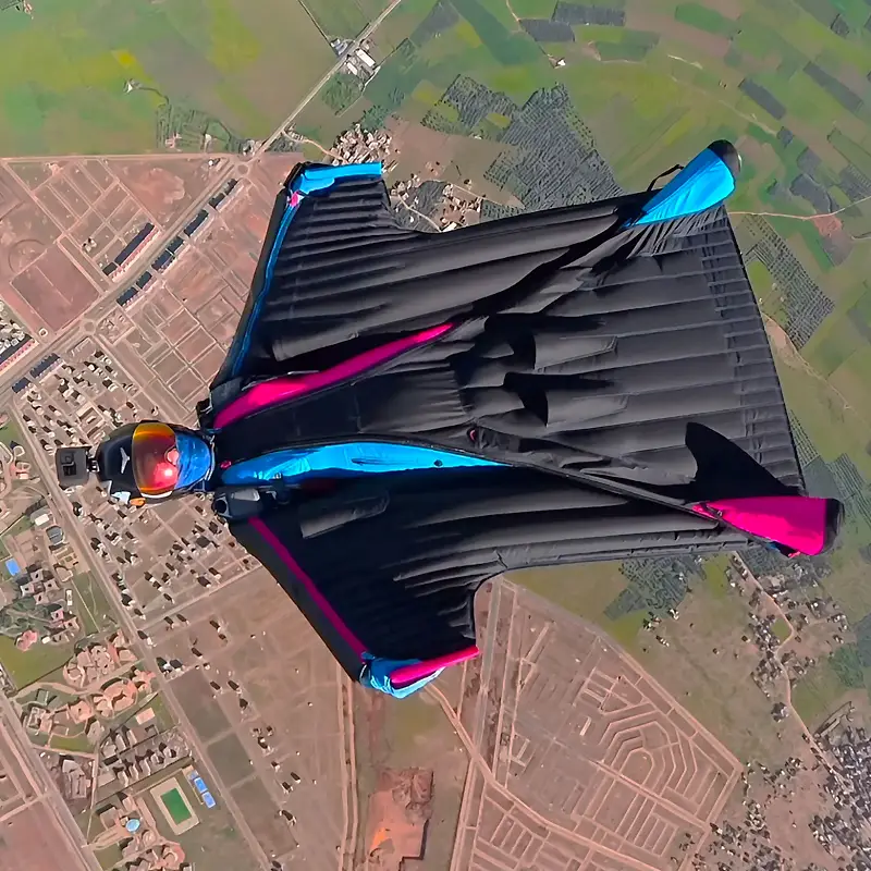 A parachutist is back flying in an Airglide Inspire 3 wingsuit. The suit is all black with pink and blue touches.